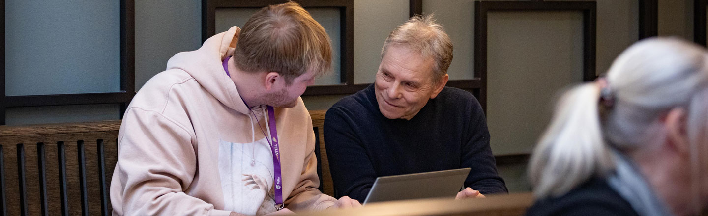 Ung mann og eldre mann smiler til hverandre, sittende ved et bord med en laptop stående på bordet.