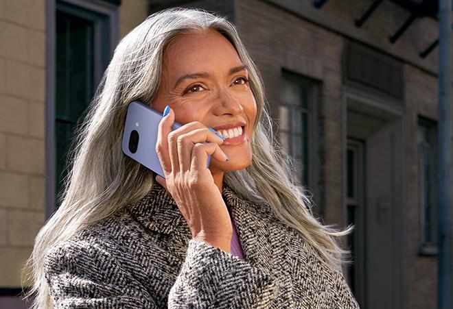 Blond, langhåret, smilende kvinne iført kåpe smiler og holder en lavendelfarget Pixel 9a opp mot øret.
