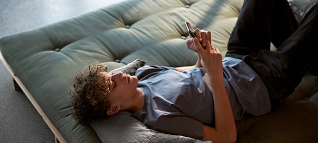 Ungdomsgutt ligger på en divan og ser opp mot mobilen sin.