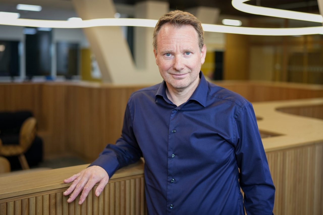 Man with blue shirt leaning against a desk at a workplace