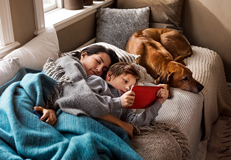 Mor, barn og hund ligger i sofaen og slapper av med mobil og nettbrett.