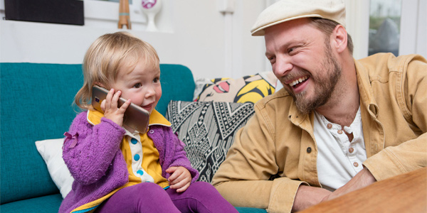 Lita jente prater energisk i mobilen. Far sitter ved siden av og ler.