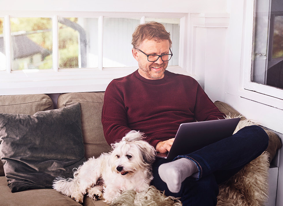 Blid mann med briller sitter og ser på laptopen i sofaen. Ved siden av ligger hans trofaste, firbeinte venn.