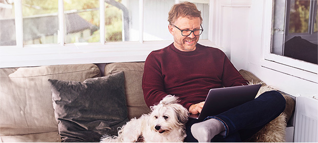 Mann sitter med laptop i sofa, en hund med gøyal frisyre ligger ved hans side.