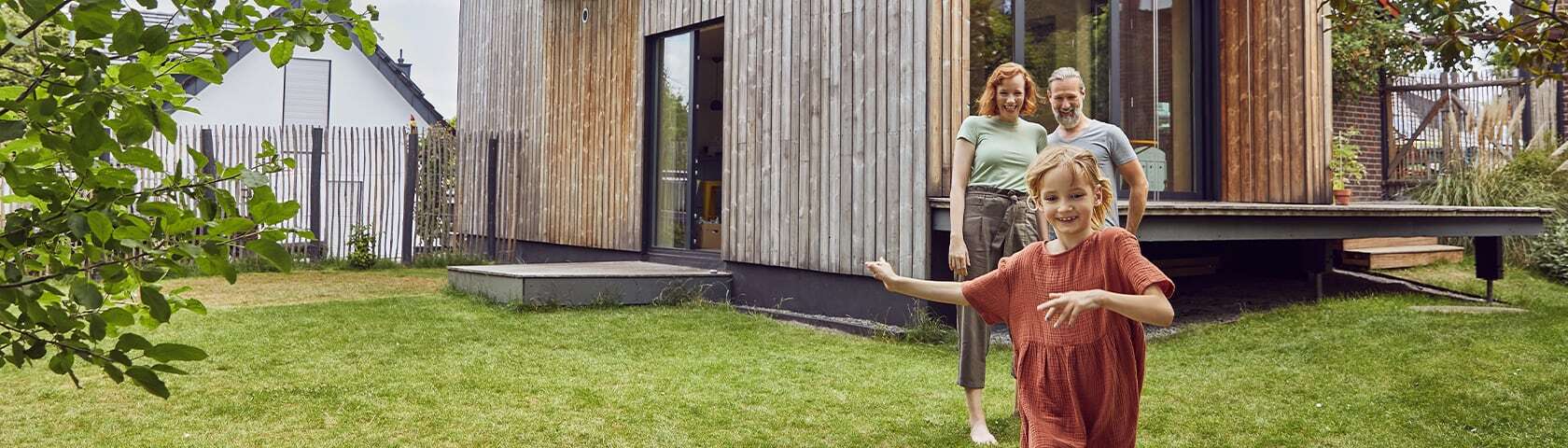 En liten familie står foran et moderne hus mens et barn løper i hagen.