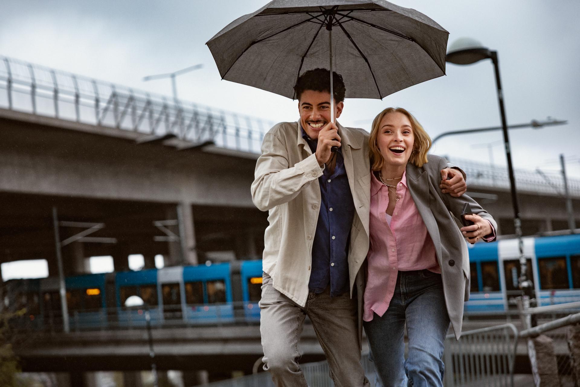 Happy couple running under an umbrella in the city, with a bridge and a train in the background.