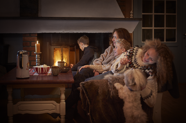 En familie sitter i en sofa foran en peis i lyset fra TVen på hytta