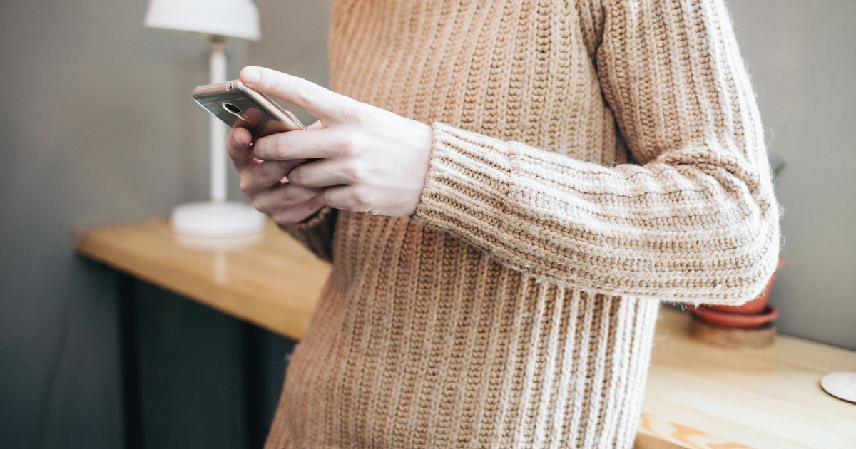 Mann iført beige strikkegenser står lent mot et bord og skriver en melding på mobilen sin. I bakgrunnen skimtes en bordlampe.