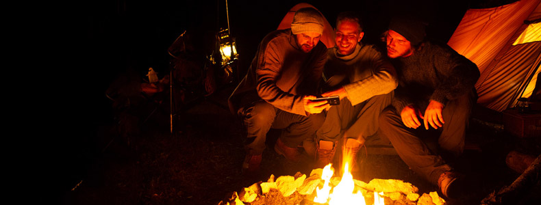 Tre kompiser sitter rundt et bål og ser på den enes mobiltelefon