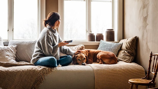 En dame holder en mobil og sitter på en sofa sammen en hund 