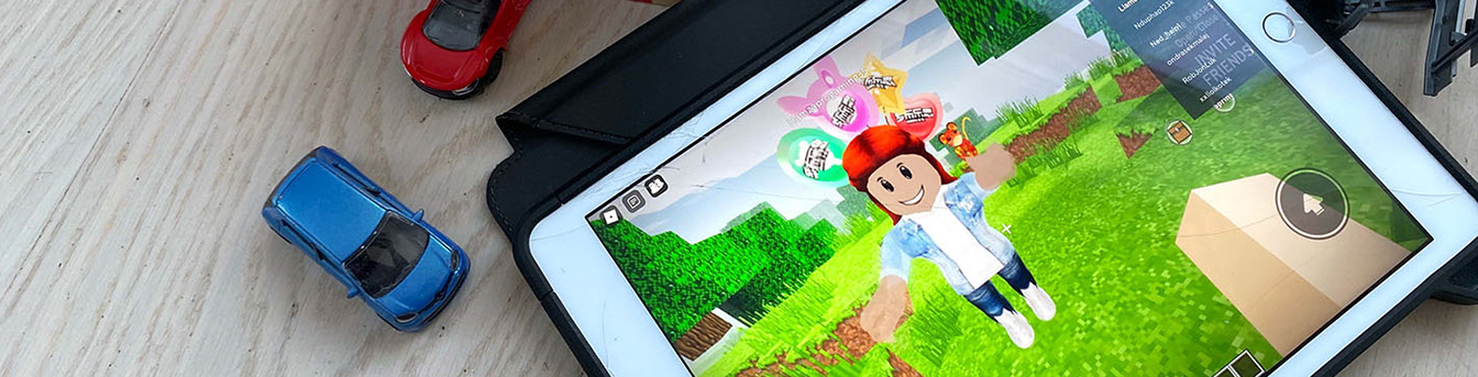 Roblox på skjermen på et nettbrett liggende på et bord, med noen lekebiler ved siden av nettbrettet.