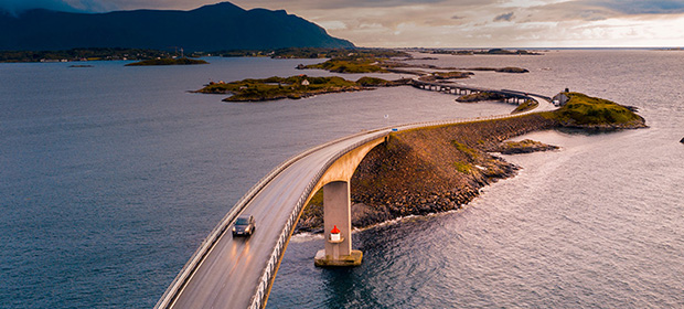 Bil kjører over en bro ute i havgapet med holmer, fjell og sjø i bakgrunnen
