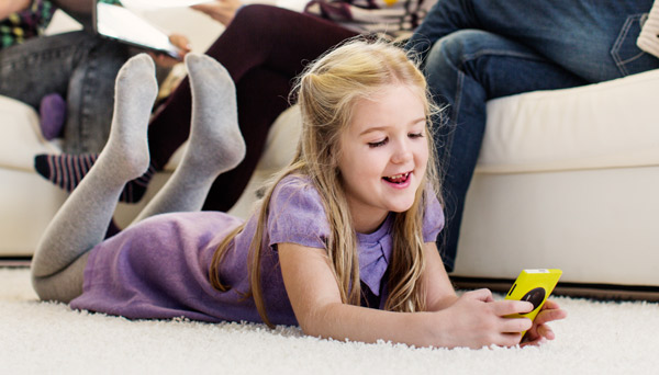 Blid jente ligger på teppet i stua og ser på mobilen. De voksne sitter i sofaen bak.