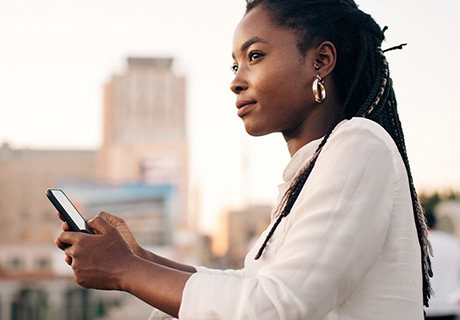 Kvinne ser utover bybildet mens hun lener seg mot et gjerde og holder mobilen i hånda.