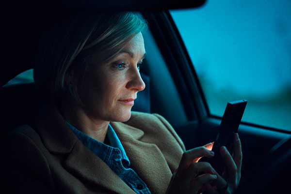 Kvinne i førersetet i parkert bil, det er skumring ute, og mørkt i bilen. Hun ser på mobilen, som delvis lyser opp ansiktet hennes.