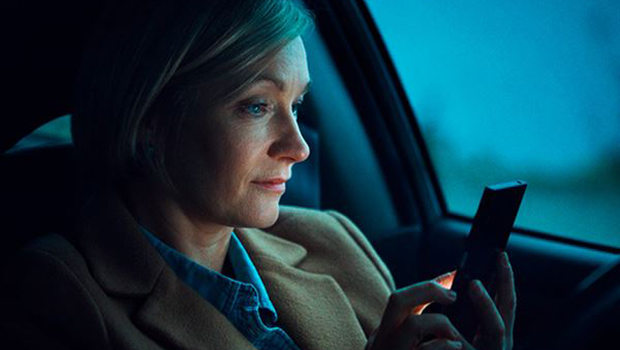 Kvinne i førersetet i parkert bil, det er skumring ute, og mørkt i bilen. Hun ser på mobilen, som delvis lyser opp ansiktet hennes.