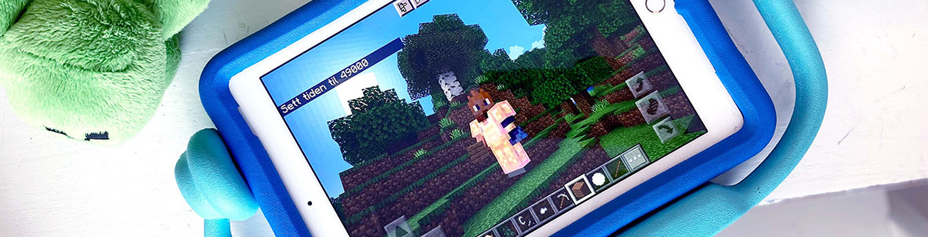 Minecraft på nettbrett liggende på et bord med en bamse stående ved siden av nettbrettet.