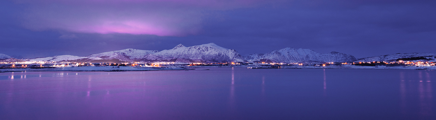 Utsikt over vinterkledd by som speiler seg i sjøen på kvelden.