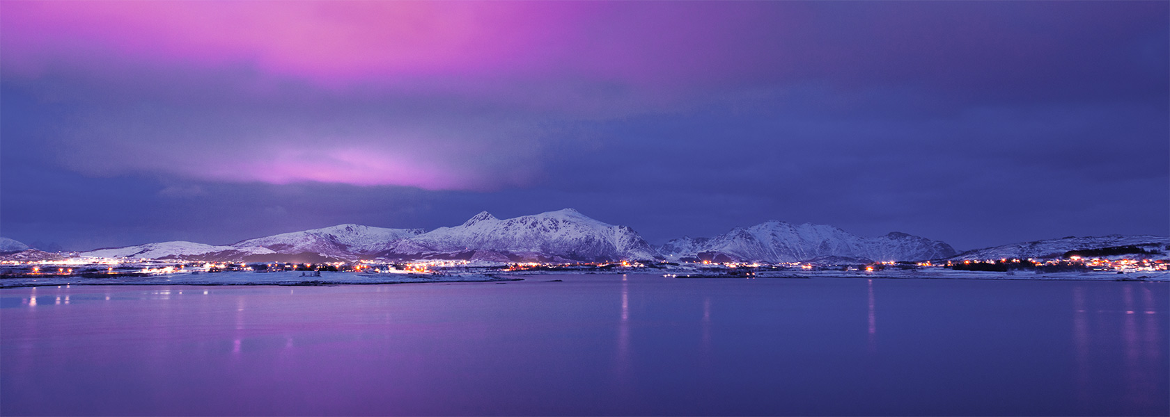Utsikt over vinterkledd by som speiler seg i fjorden på kvelden.