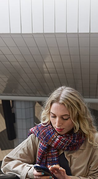 Kvinne med blondt hår og et stort skjerf står i en rulletrapp på vei opp fra t-banen og taster på mobiltelefonen