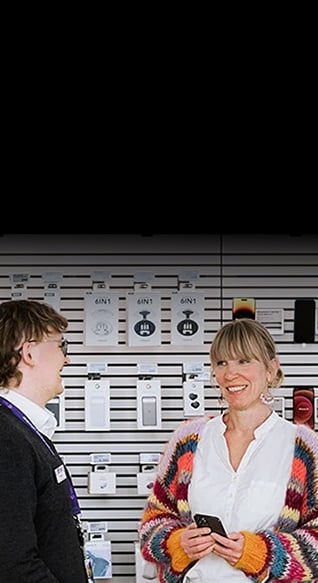 Ansatt og kunde snakker sammen i en Telia-butikk, bak dem ser vi diverse mobiler og tilbehør på veggen.