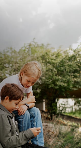 To små barn sitter ute i parkmessig opparbeidet uteområde, og ser på den ene guttens mobiltelefon.