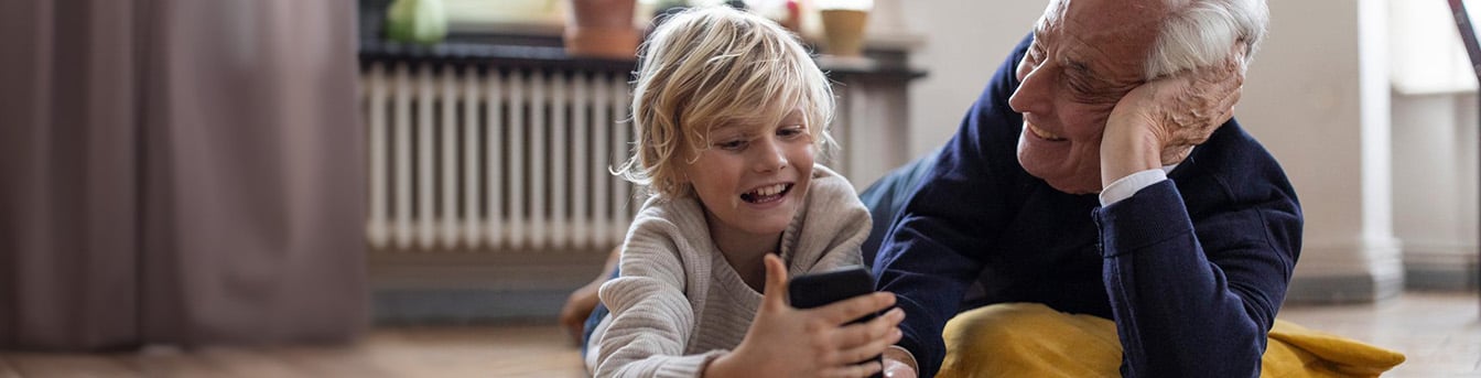 Eldre mann med grått hår og mørk genser ligger på gulvet med en gul pute, og smiler til en liten gutt med blondt hår mens de ser på mobiltelefonen