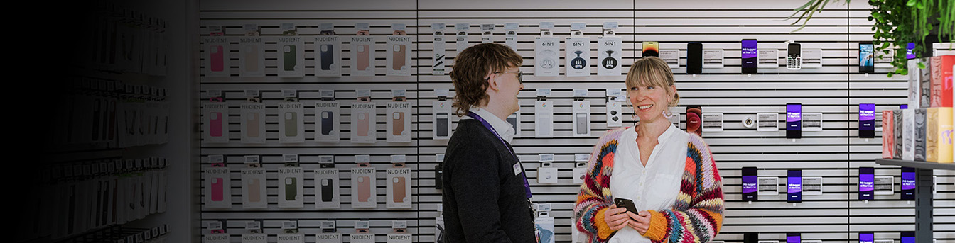 Ansatt og kunde snakker sammen i en Telia-butikk, bak dem ser vi diverse mobiler og tilbehør på veggen.