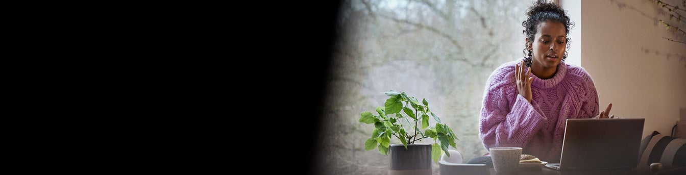 Kvinne iført ullgenser sitter ved kjøkkenbordet med kaffekopp og laptop.