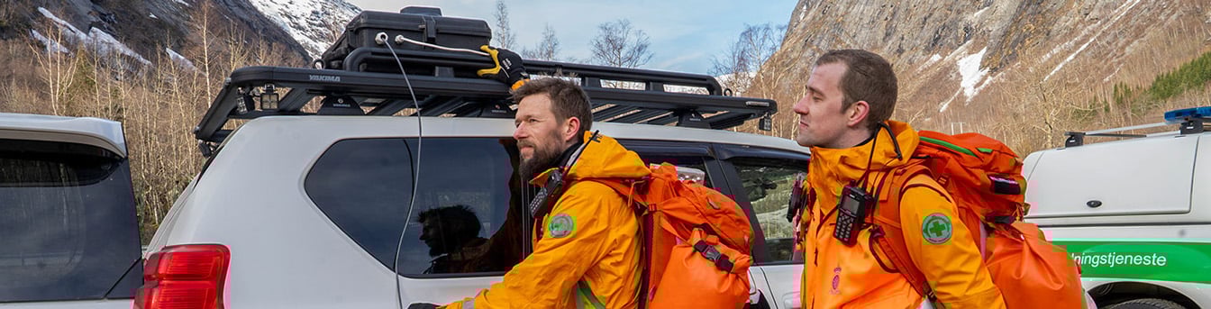 Personer fra Norsk Folkehjelp ute i felten med utstyr og biler, fjellandskap ses i bakgrunnen.