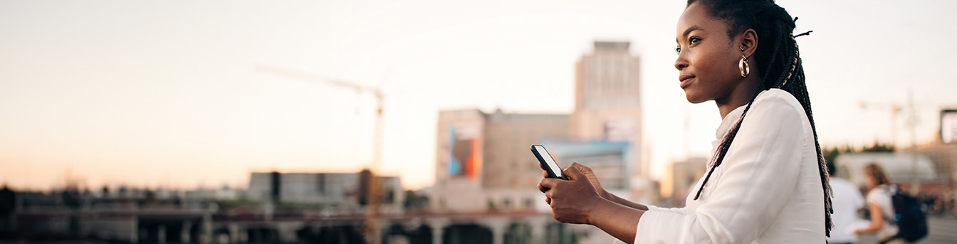 Ung kvinne holder mobilen mens hun lener seg på et gjerde med bymiljø i bakgrunnen.
