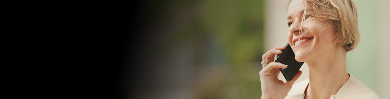 Smilende, korthåret blond kvinne, sett fra siden, står ute i bymiljø og snakker i mobilen.