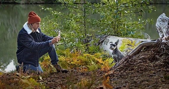 Mann på tur med hunden i skogen ved et tjern. De har stoppet på et frodig sted for å få et godt portrett av den firbeinte ute i naturen. Hunden sitter rolig.