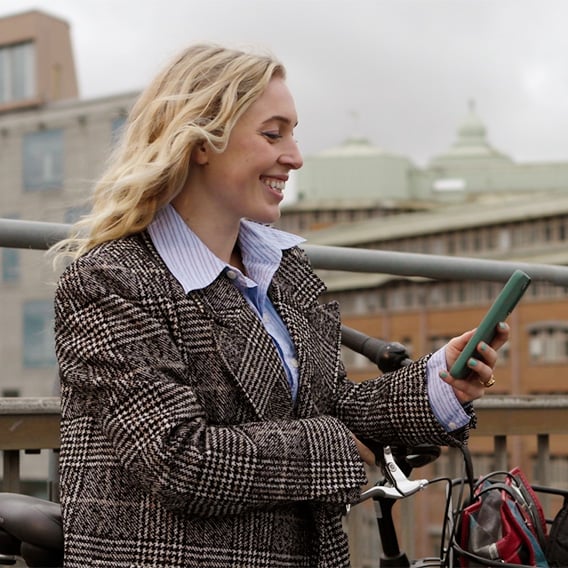 Ung kvinne i kåpe står på en bro i byen med sykkelen sin og ser på mobilen mens hun smiler.