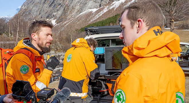 Personer fra Norsk Folkehjelp ute i felten med utstyr og biler, fjellandskap ses i bakgrunnen.