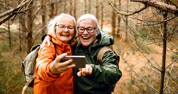 To eldre kvinner på tur i skogen, de tar selfie med mobilen og smiler.