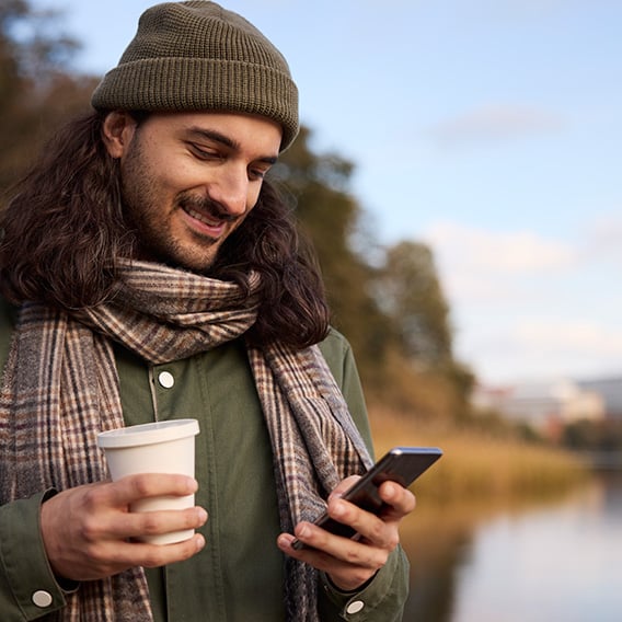 Ung mann står i høstklær ved et vann. Han smiler mens han holder mobilen i ene hånda og takeaway kaffekopp i den andre.