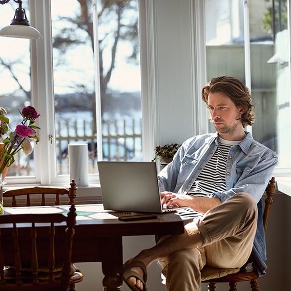 Mann sittende ved spisebord foran laptop med smart wifi router i vinduskarmen Mann sittende ved spisebord foran laptop med smart wifi router i vinduskarmen