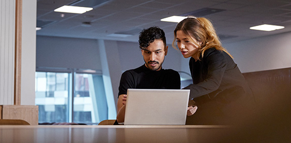 Mann og kvinne ved laptop i møterom. Mannen sitter ved pulten, kvinnen står ved siden av han. Begge ser på skjermen på laptopen. Mann og kvinne ved laptop i møterom. Mannen sitter ved pulten, kvinnen står ved siden av han. Begge ser på skjermen på laptopen.