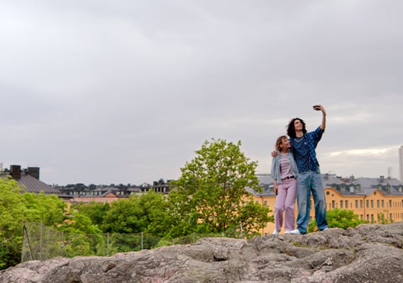 To personer står og tar selfie på noen svaberg, vi ser bymiljø i bakgrunnen.
