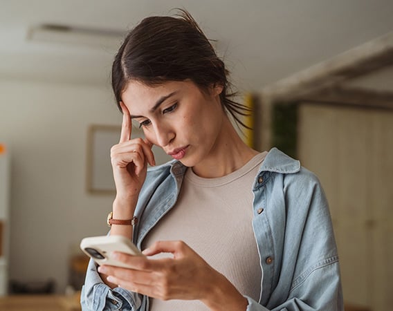 Ung kvinne står i stua og ser på mobilen med et litt usikkert ansiktsuttrykk.