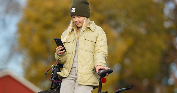 Jente kledd i jakke og lue, står med sykkelen sin og ser på mobilen.