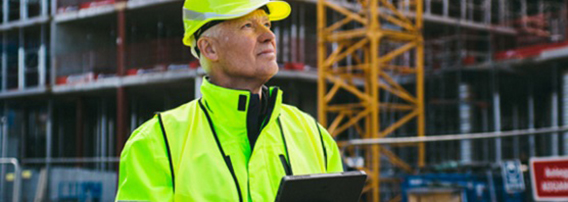 Mann iført hjelm og arbeidsklær betjener fjernkontroll til en drone som inspiserer en byggeplass