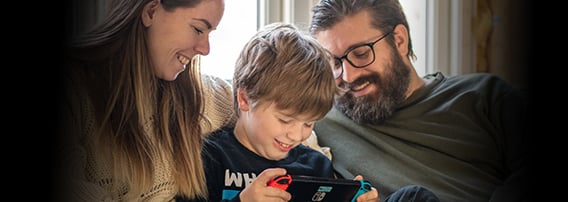 Mor, far og liten sønn sitter sammen i sofaen og smiler og ler. Sønnen holder en Nintendo Switch.