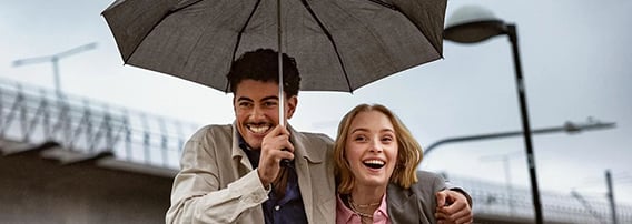Ungt, smilende par, under en paraply som han holder. Ute i byen, vi ser en trikk i bakgrunnen.