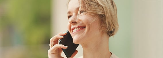 Smilende, korthåret blond kvinne, sett fra siden, står ute i bymiljø og snakker i mobilen.
