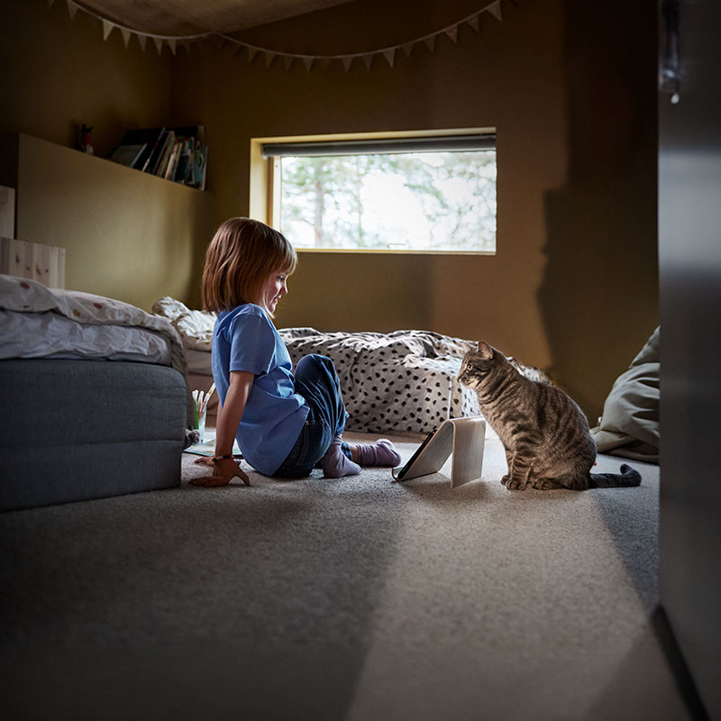 Liten jente og en katt sitter på gulvet på soverommet og ser på hverandre. Mellom dem står et nettbrett.