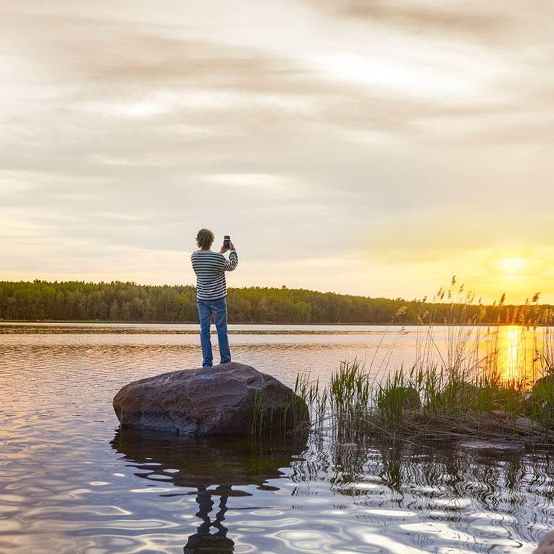Ung mann sett bakfra stående på stein i sjø, mens han tar bilde av solnedgangen med mobilen, en vakker kveld.