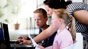 To barn og en kvinne sitter / står foran en PC. To barn og en kvinne sitter / står foran en PC.
