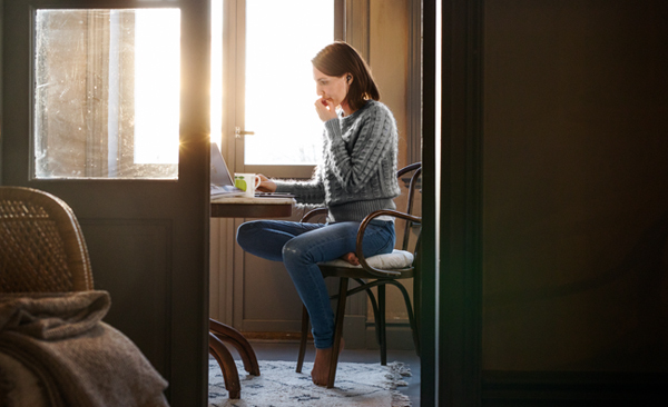Kvinne tar seg undrende til haken foran et bord med kaffekopp og laptop. Solskinn fra vinduet i bakgrunnen.